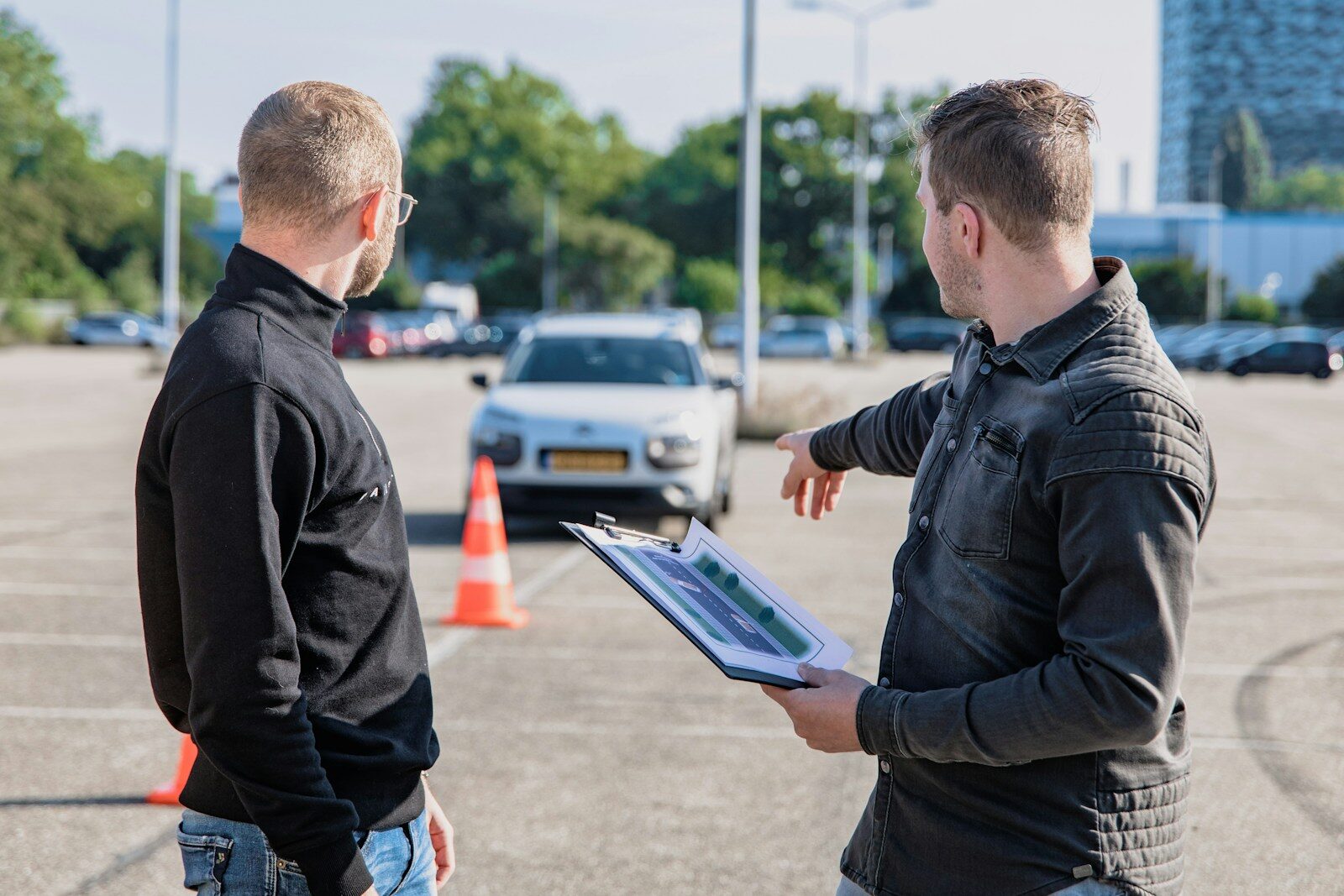 A man pointing toward a car while holding a clipboard for a driving test
